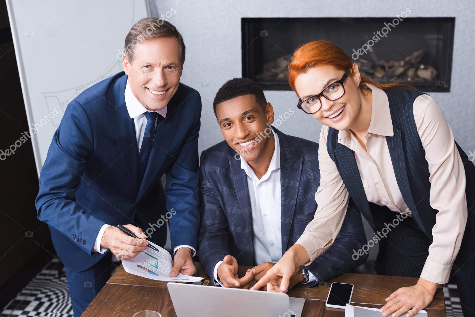 Happy multicultural businesspeople looking at camera near workplace with digital devices